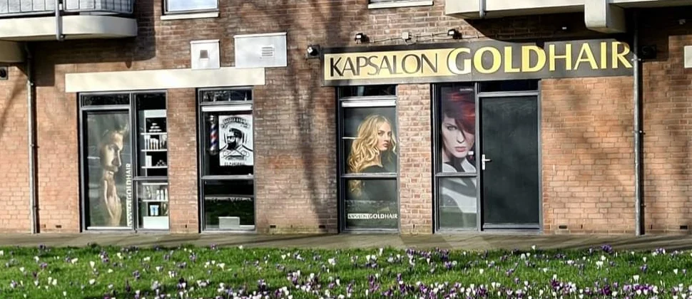 De gevel van Kapsalon Goldhair aan de Melkkelder: het zwarte bord met de gouden letters boven de etalages, krokussen in het gras